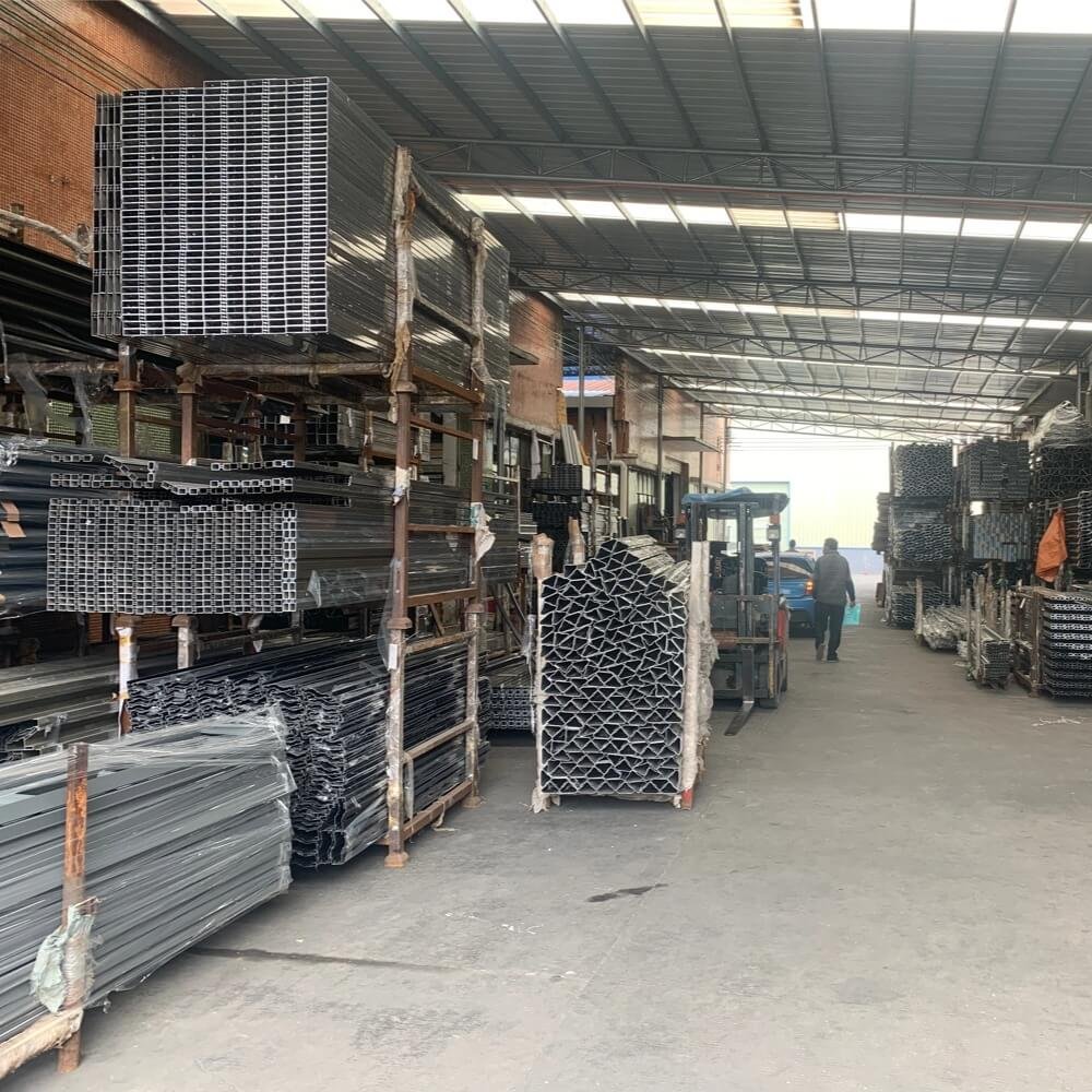 Interior view of a large-scale aluminum manufacturing facility showing massive racks of powder-coated pergola profiles and a forklift operating in the warehouse.