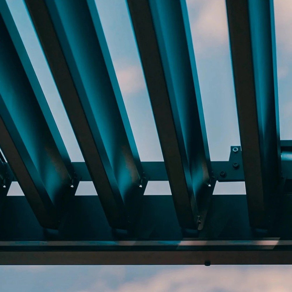 Close-up view of open, adjustable aluminum louver blades on a bioclimatic pergola against a blue sky, showcasing the precision rotation mechanism.
