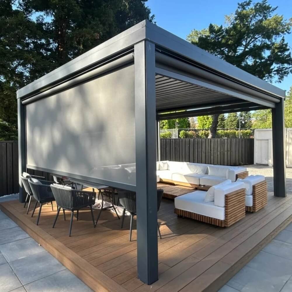 Motorized grey zip screen blinds partially lowered on an aluminum pergola, providing wind resistance and privacy for a luxury outdoor seating area.