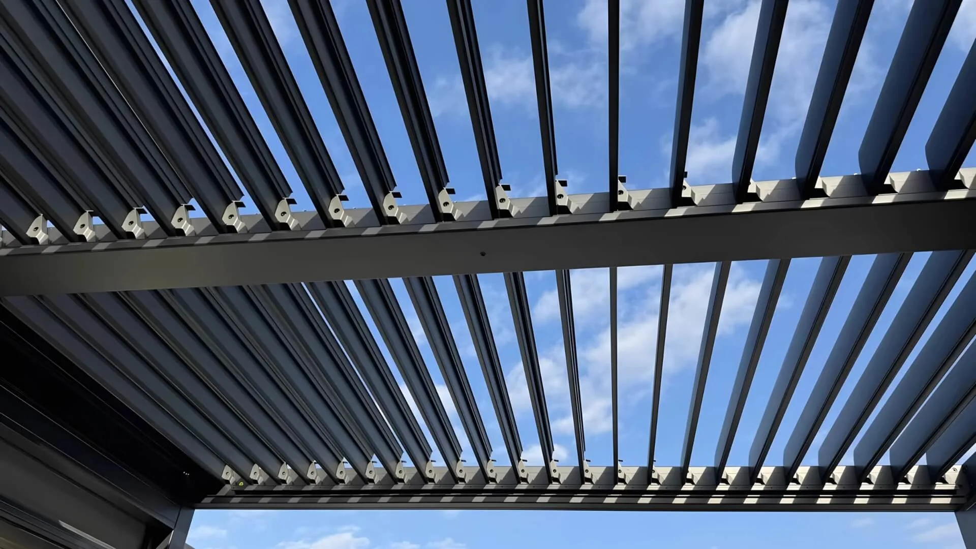Close-up view looking up at the open louvers of a dark grey aluminum bioclimatic pergola against a blue sky, showcasing the precision engineering and internal mechanisms of the adjustable roof system.