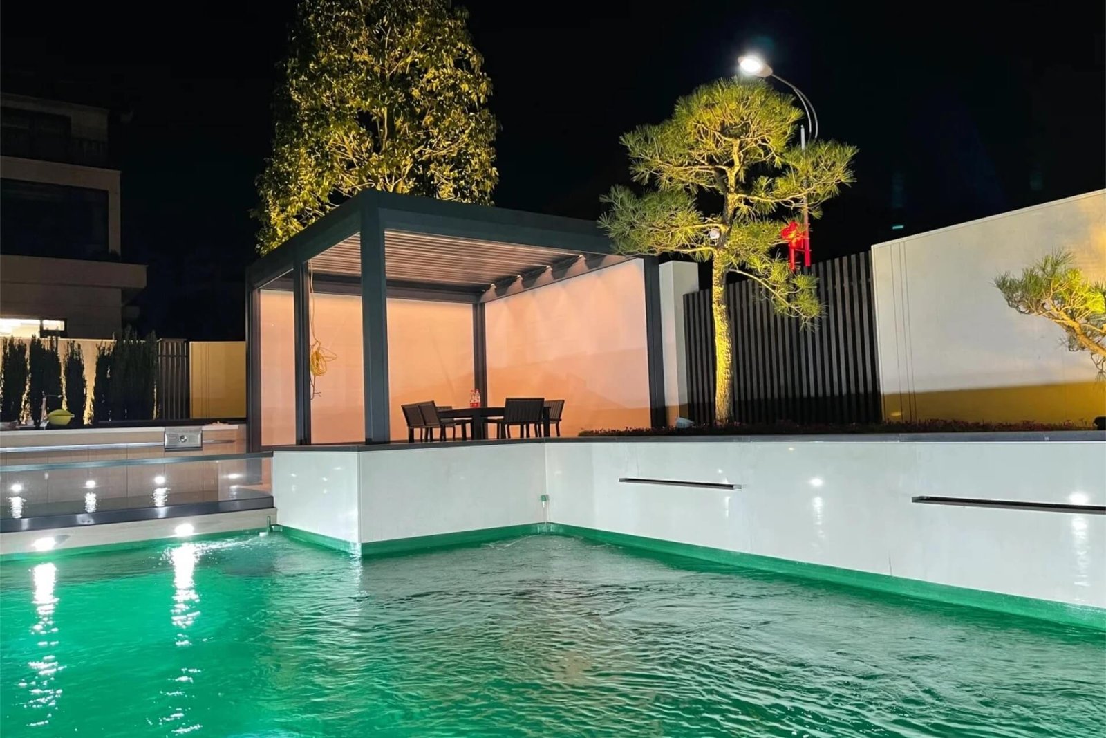 Stunning night view of a custom dark grey aluminum pergola next to a swimming pool in Penang, featuring integrated LED lighting and white privacy screens.