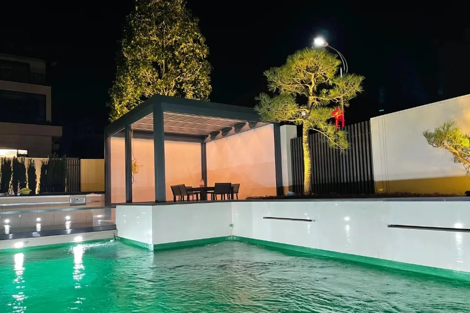 Stunning night view of a custom dark grey aluminum pergola next to a swimming pool in Penang, featuring integrated LED lighting and white privacy screens.