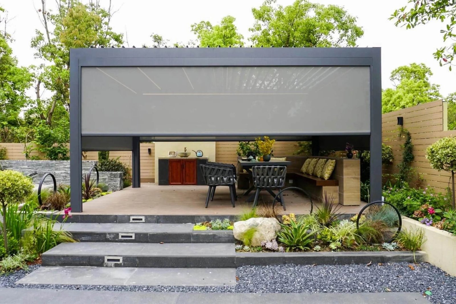 Premium dark grey freestanding aluminum bioclimatic pergola with partially lowered zip screens, sheltering a wooden deck and outdoor kitchen area in a modern landscaped garden.