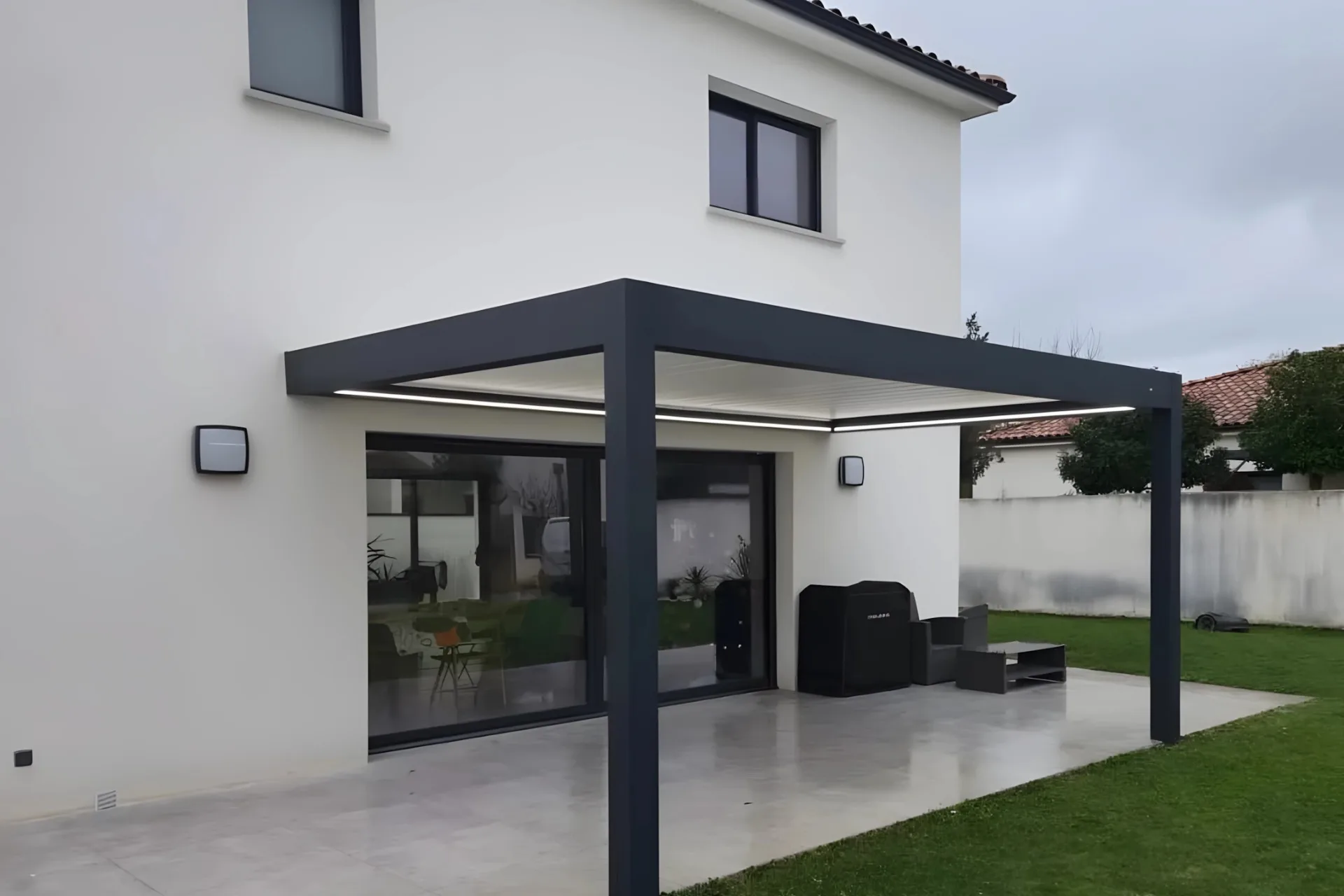 An anthracite grey wall-mounted bioclimatic aluminum pergola installed over a wooden deck patio next to a modern house, showing the seamless indoor-outdoor living space extension.