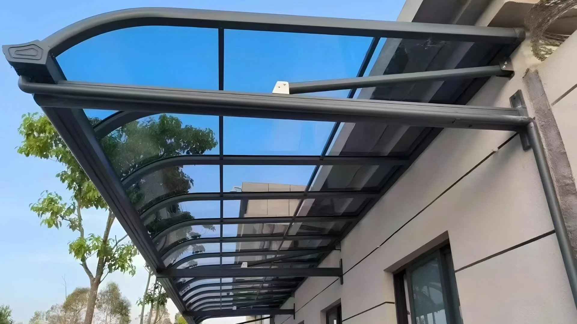 A custom wall-mounted aluminum patio cover featuring UV-blocking blue polycarbonate roof panels, installed over a building entryway to provide all-weather protection.