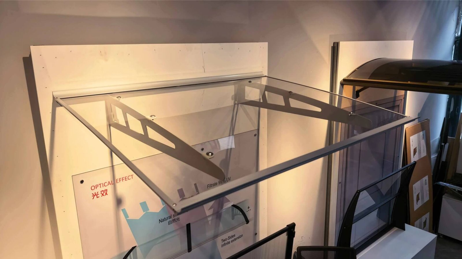 A factory showroom display of a modern slant roof polycarbonate door canopy, highlighting its clear optical effect, 99% UV filtration, and heavy-duty aluminum bracket structure.