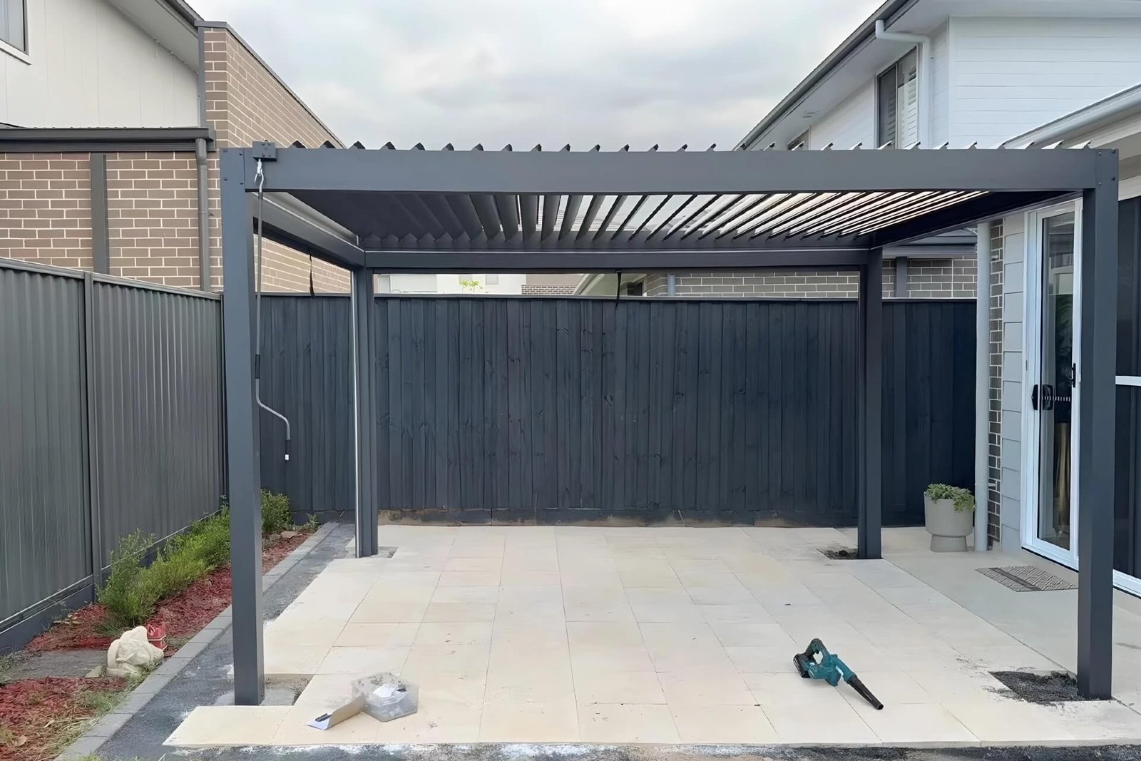 Real-world installation of a dark grey manual hand-crank aluminum pergola on a residential backyard patio, featuring adjustable louvers for natural ventilation.