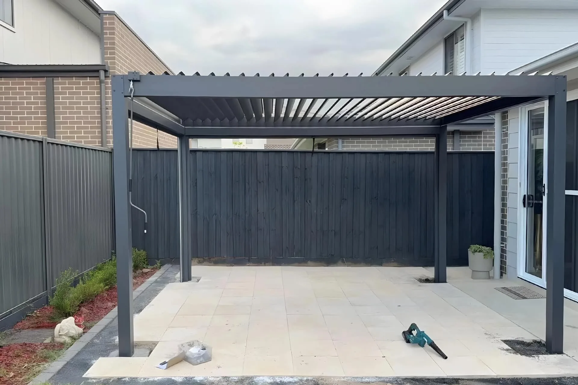 Real-world installation of a dark grey manual hand-crank aluminum pergola on a residential backyard patio, featuring adjustable louvers for natural ventilation.